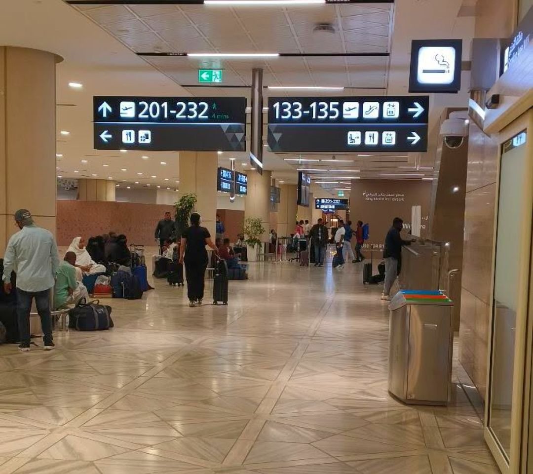 Saguão de desembarque de aeroporto em Riade, com placas de embarque e passageiros com malas