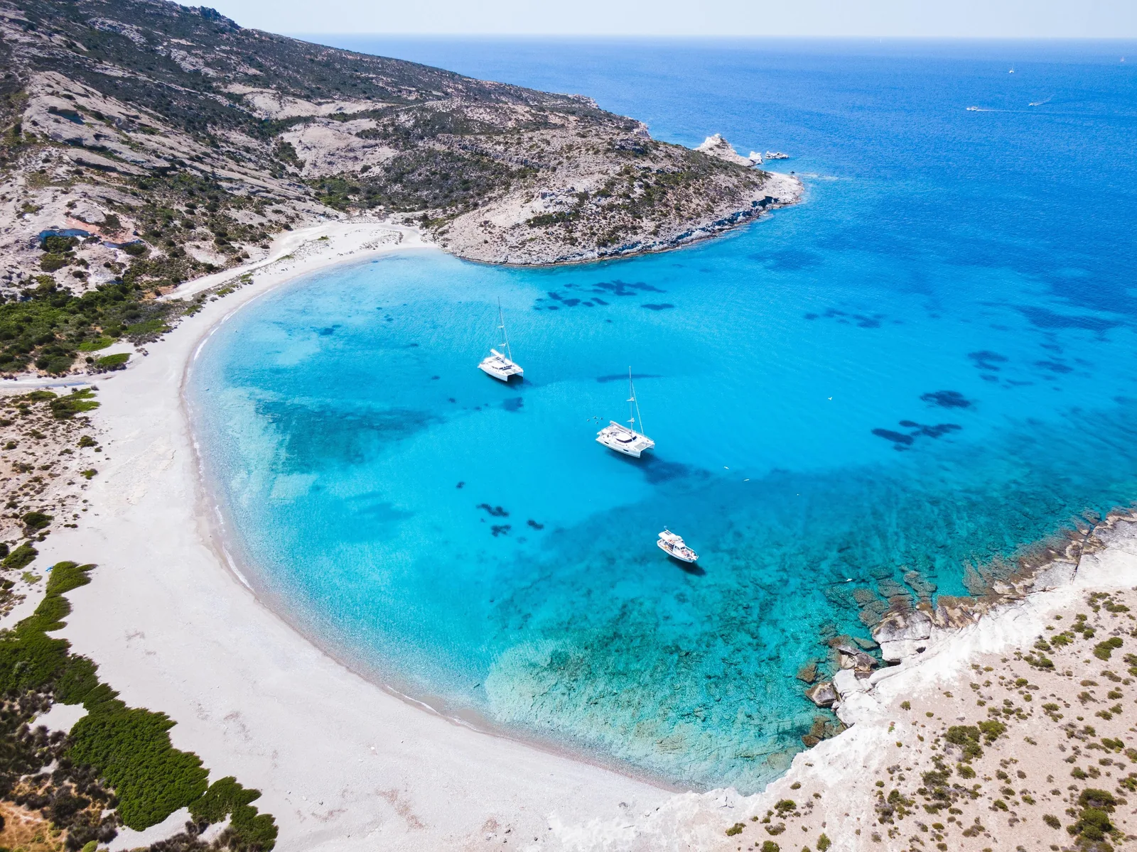 Enseada de areia clara e mar turquesa na costa de Milos, vista do alto, com veleiros ancorados