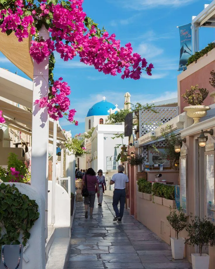 Visitantes caminhando por uma ruela de Oia, em Santorini, com buganvílias e uma igreja de cúpula azul ao fundo