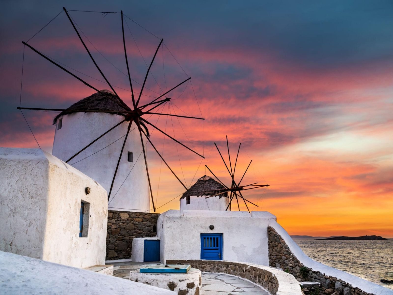 Pôr do sol com o céu alaranjado atrás dos moinhos de vento de Mykonos, junto ao mar