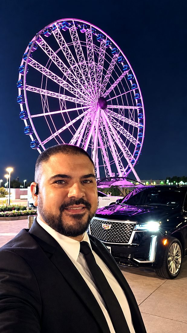 Alisson ao lado da SUV, com a roda-gigante iluminada ao fundo em Dallas
