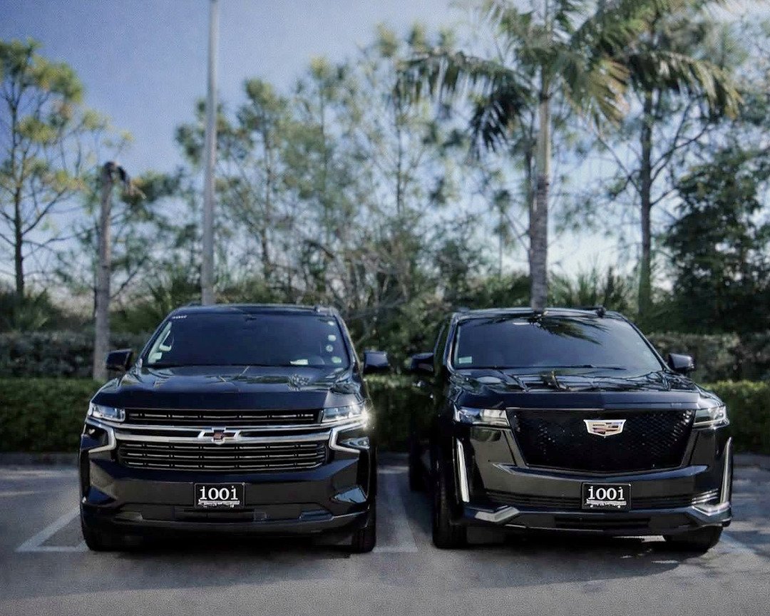 Duas SUVs pretas da 1001 Transportation, uma Chevrolet e uma Cadillac, estacionadas lado a lado entre palmeiras