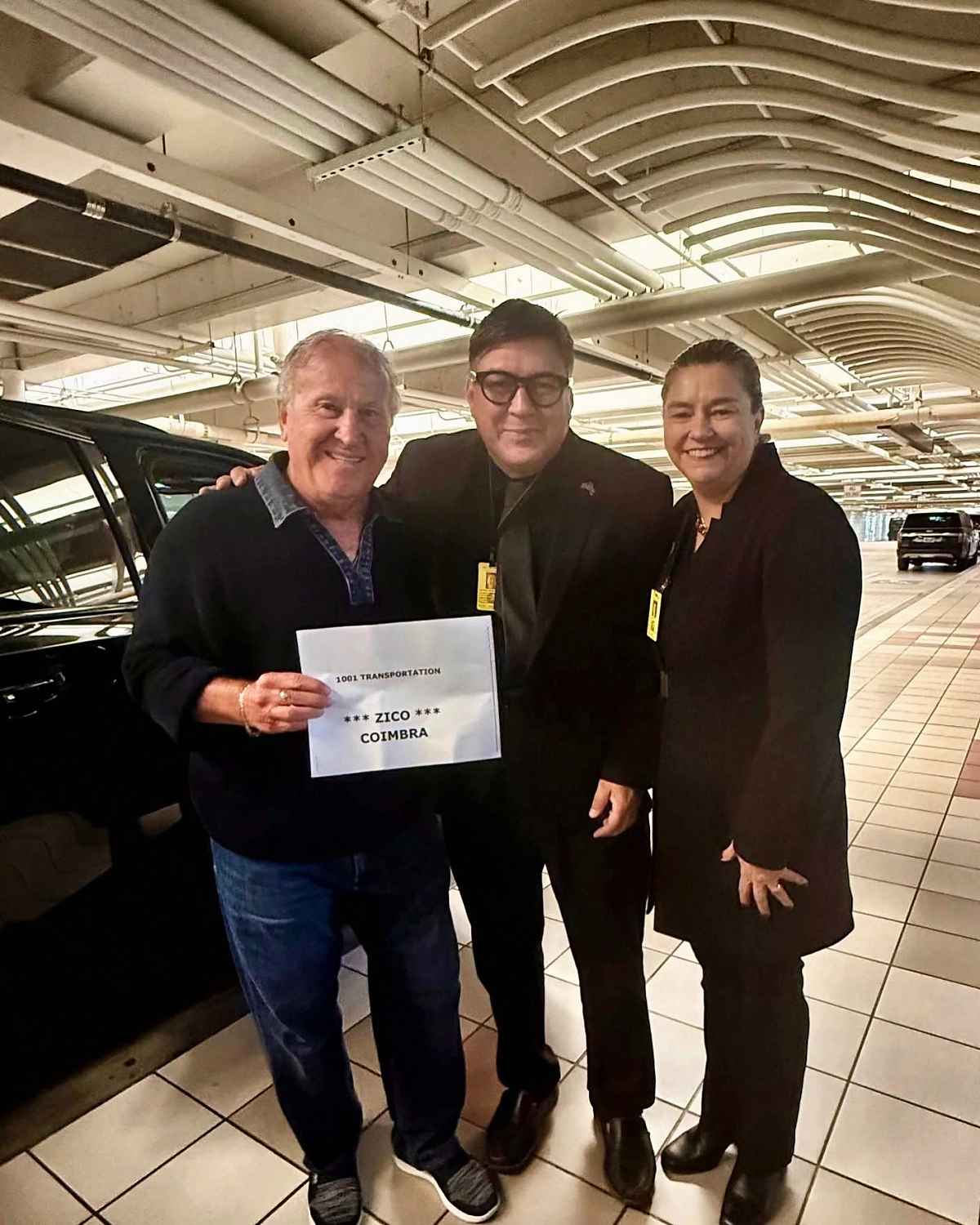Zico ao lado de Marcelo e Débora, da 1001 Transportation, segurando a placa de recepção no aeroporto