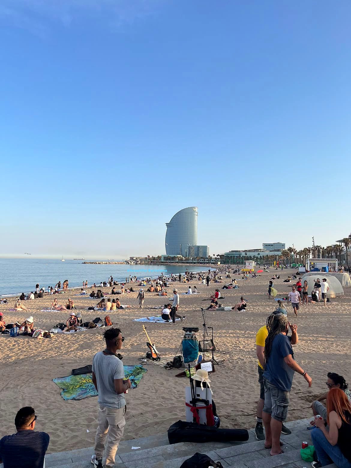 Praia da Barceloneta em Barcelona no fim da tarde, com músicos na areia e o Hotel W em forma de vela ao fundo