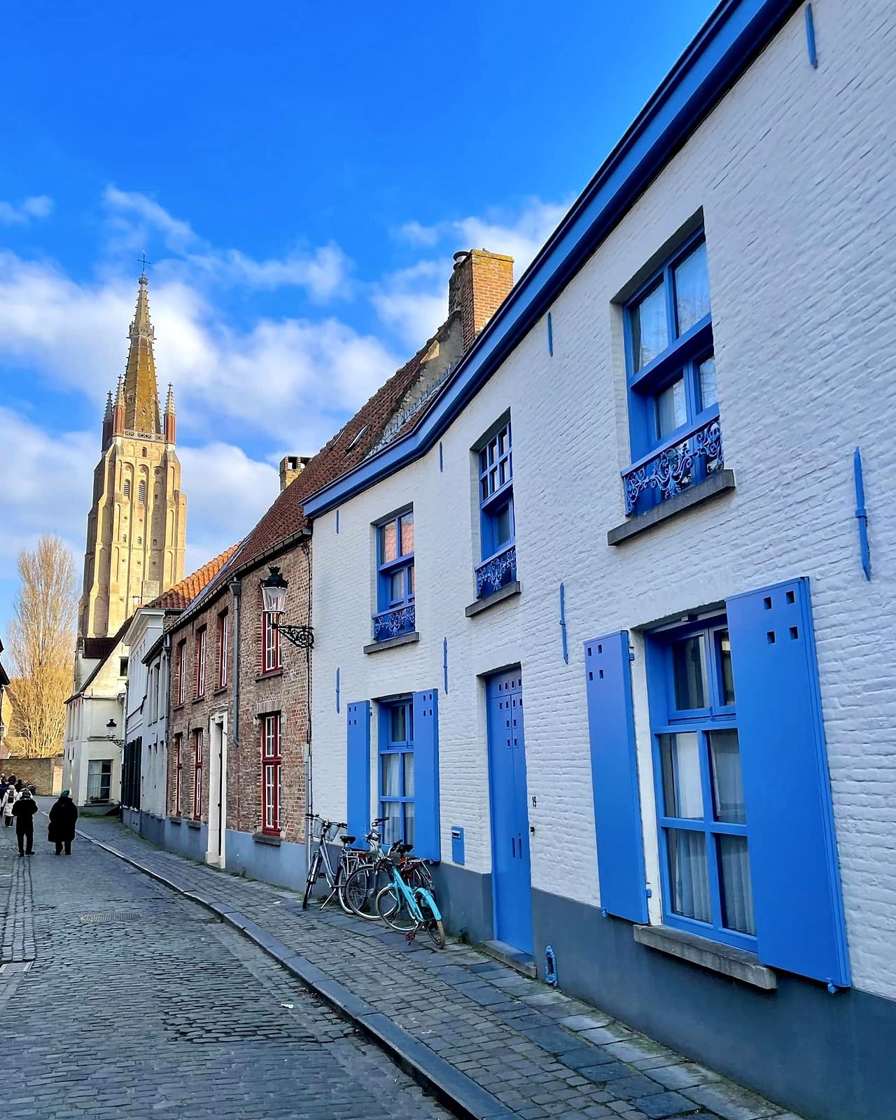 Rua de paralelepípedos em Bruges com casas de janelas azuis e a torre da Igreja de Nossa Senhora ao fundo