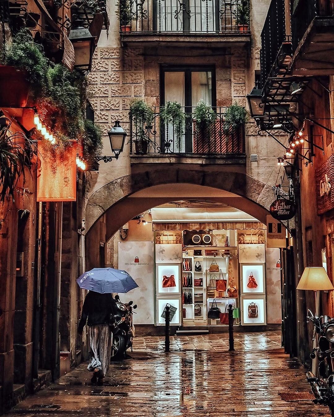 Rua estreita de El Born em Barcelona com arco de pedra, lojas e luzes, em dia de chuva