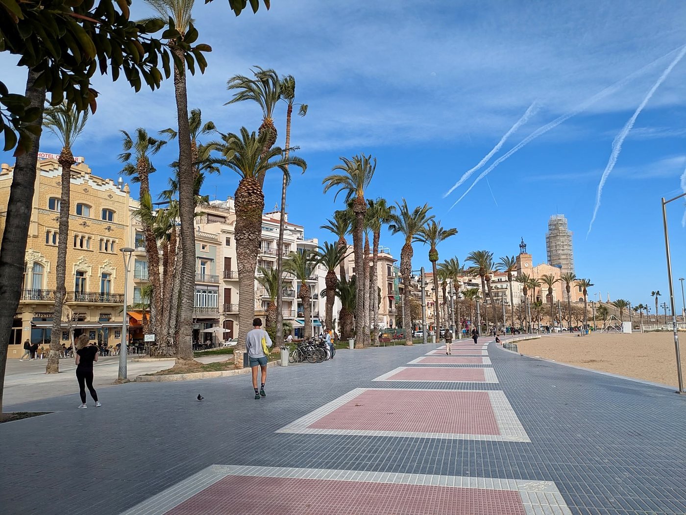 Calçadão de Sitges com palmeiras, prédios históricos, a praia e a igreja de Sant Bartomeu i Santa Tecla