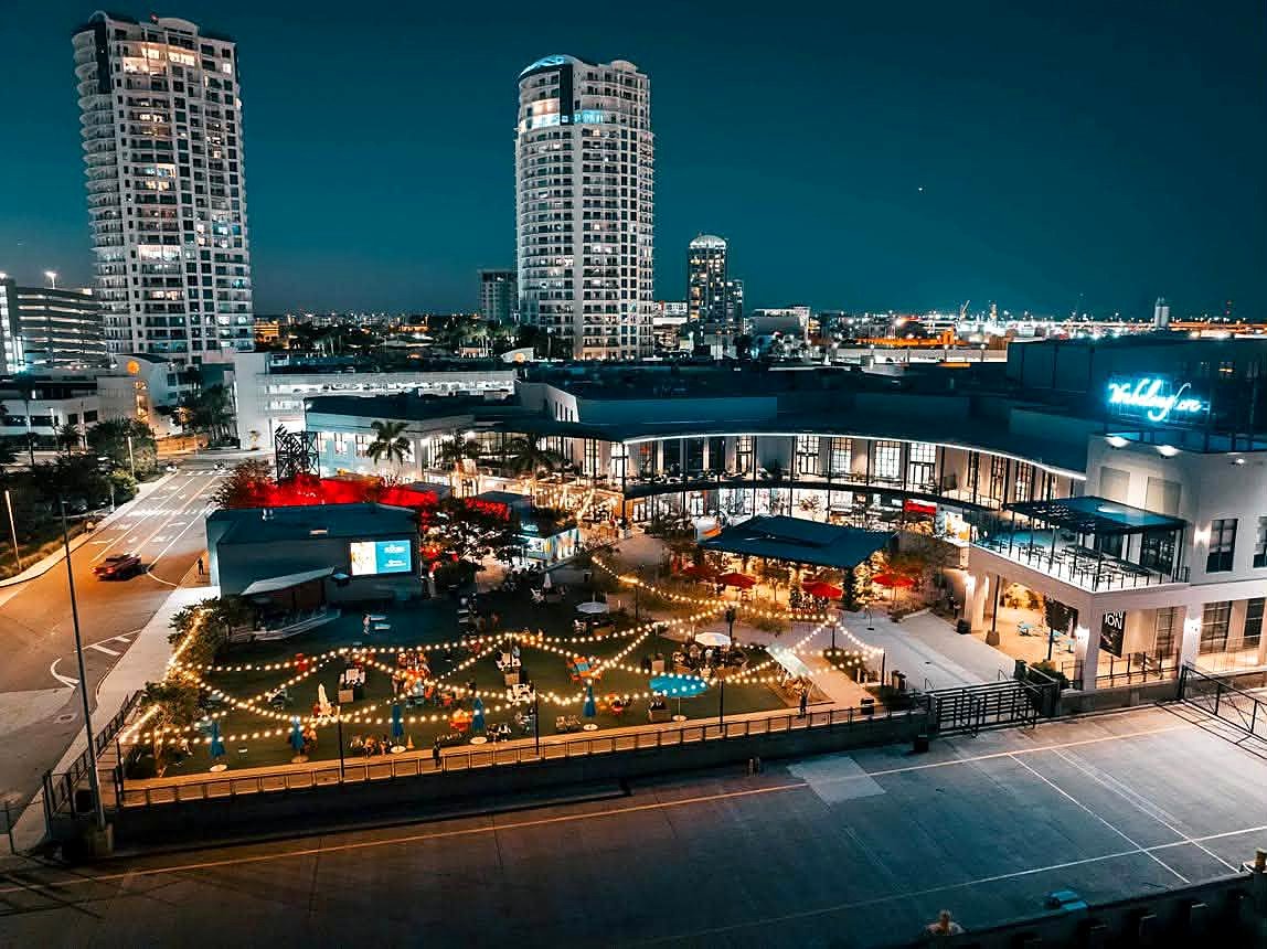 Sparkman Wharf iluminado à noite, com gramado, mesas e varal de luzes no centro de Tampa