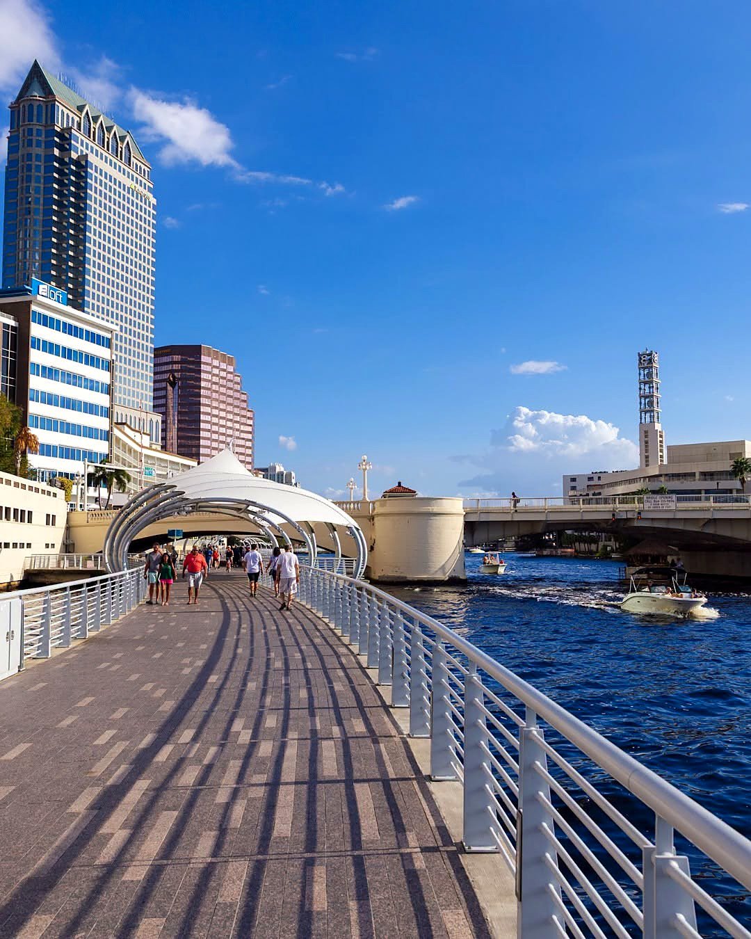 Calçadão do Tampa Riverwalk à beira do rio Hillsborough, com prédios do centro e barcos