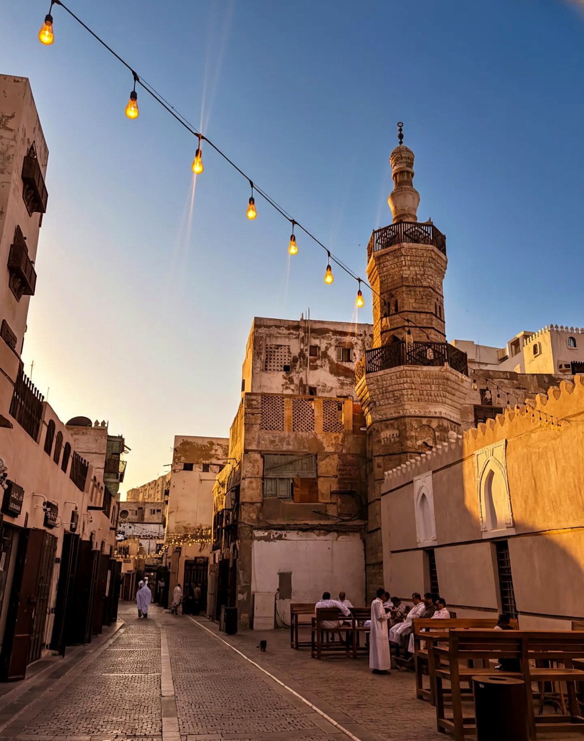 Rua de Al-Balad ao entardecer com minarete de pedra e varal de lâmpadas