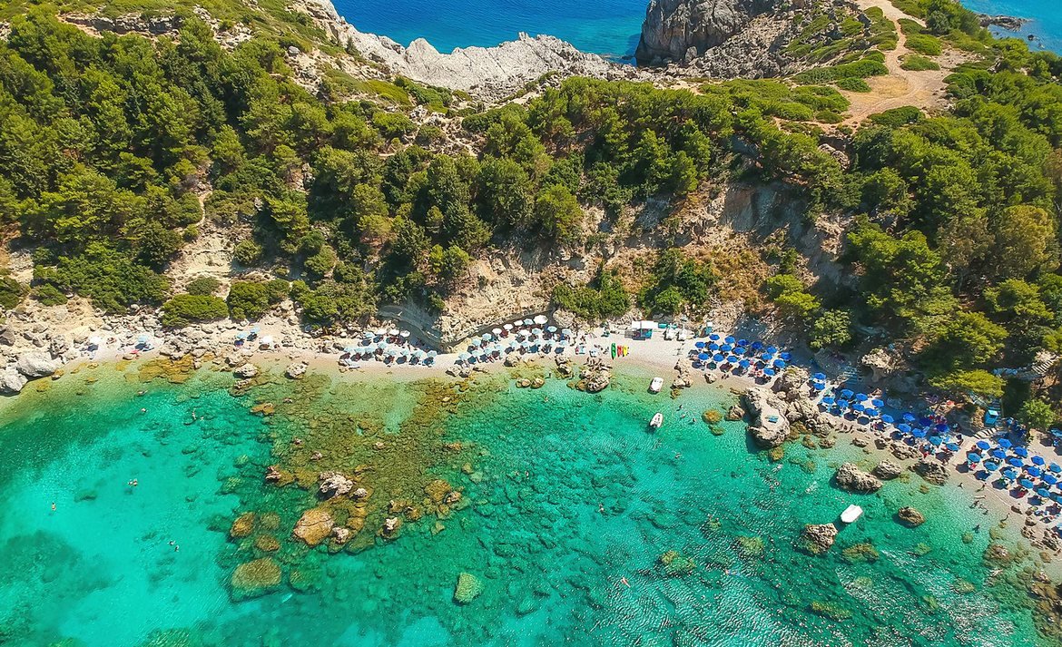 Faixa de praia de Anthony Quinn Bay vista de cima, com guarda-sóis e mar transparente