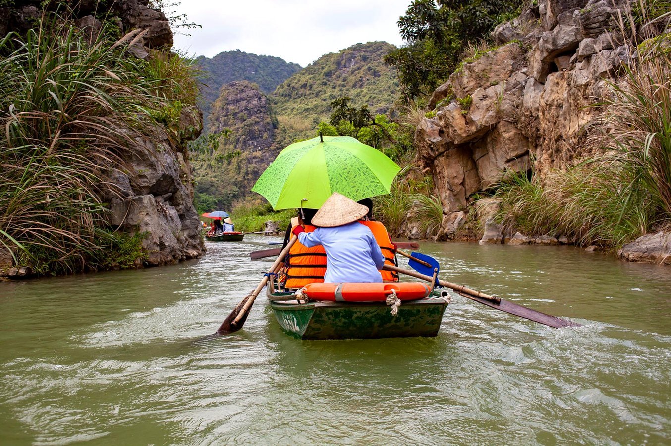 Remadora com chap&eacute;u c&ocirc;nico conduzindo turistas sob guarda-chuva verde em rio de Ninh Binh