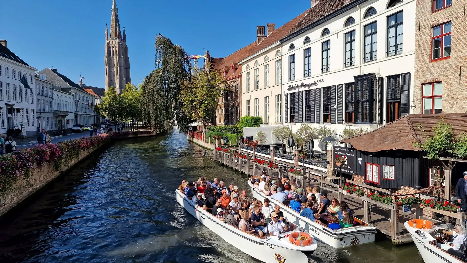 Barcos turísticos navegando pelo canal Dijver, em Bruges, com a torre da Igreja de Nossa Senhora ao fundo