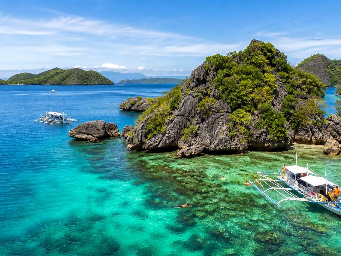 Barcos bangka e pessoas fazendo snorkel ao lado de ilhota de pedra em passeio de island hopping nas Filipinas