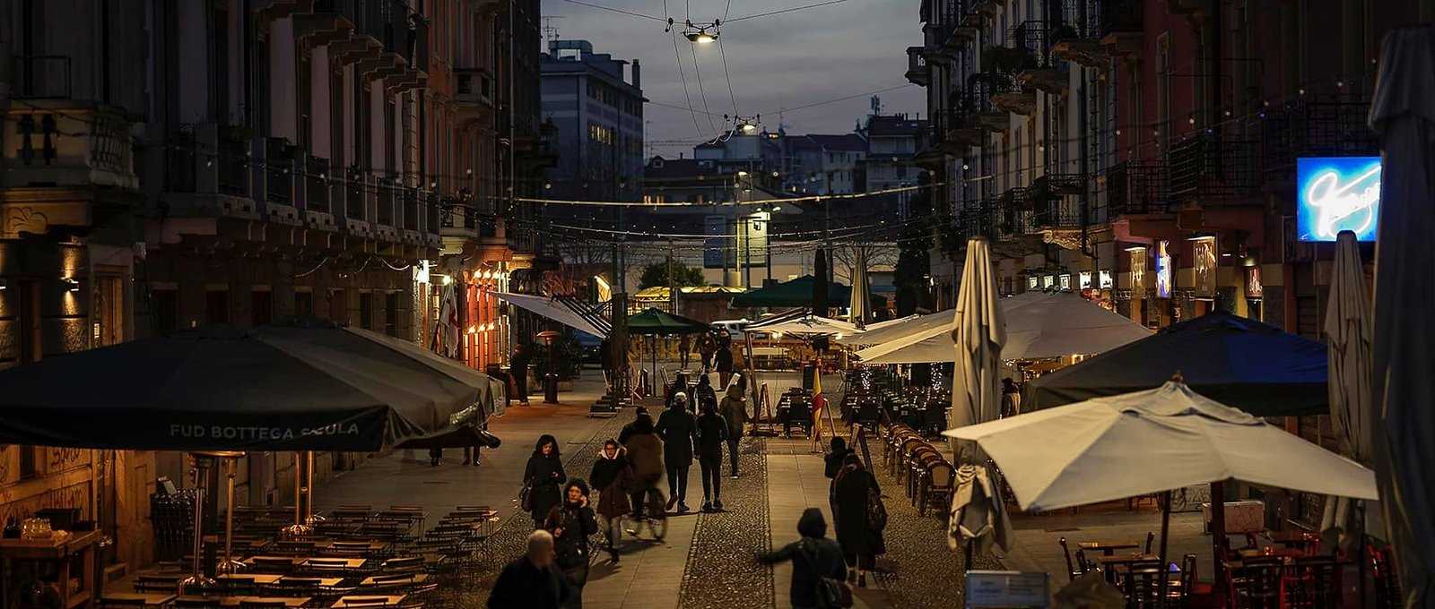 Rua de pedestres em Milão ao anoitecer com cafés, guarda-sóis e luzes penduradas
