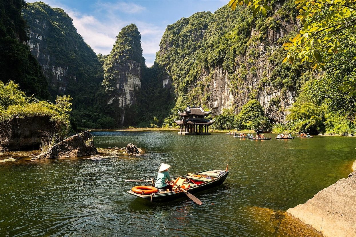 Remadora com chap&eacute;u c&ocirc;nico em barco no rio de Trang An, em Ninh Binh, com pavilh&atilde;o e montanhas de calc&aacute;rio