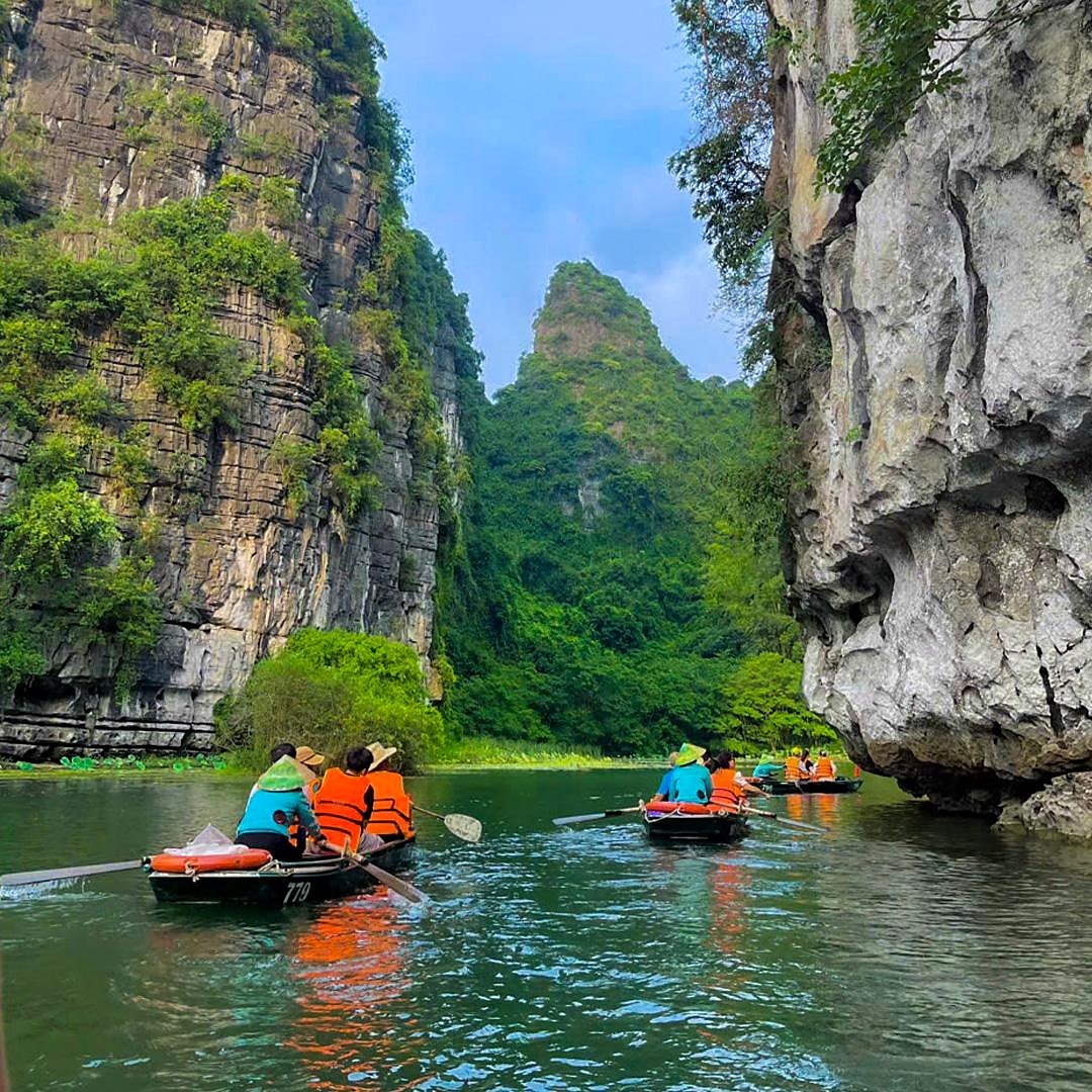 Turistas com coletes laranja em barcos a remo entre pared&otilde;es de calc&aacute;rio em Trang An, Ninh Binh