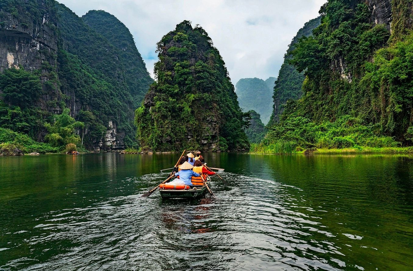 Barco a remo com turistas no rio de Trang An, cercado por montanhas de calc&aacute;rio cobertas de mata