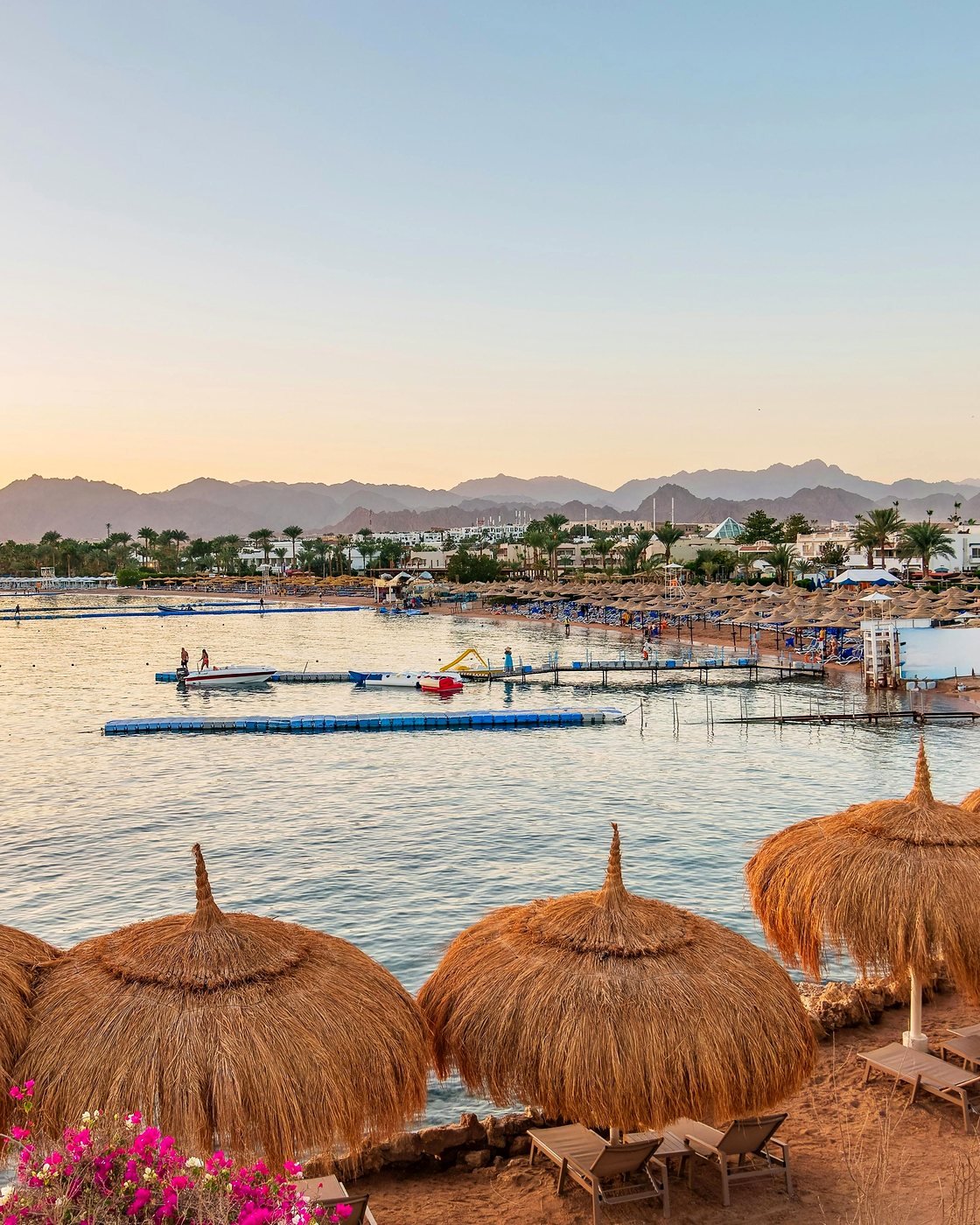 Praia de resort com guarda-sóis de palha em Sharm el-Sheikh ao entardecer, com as montanhas do Sinai ao fundo