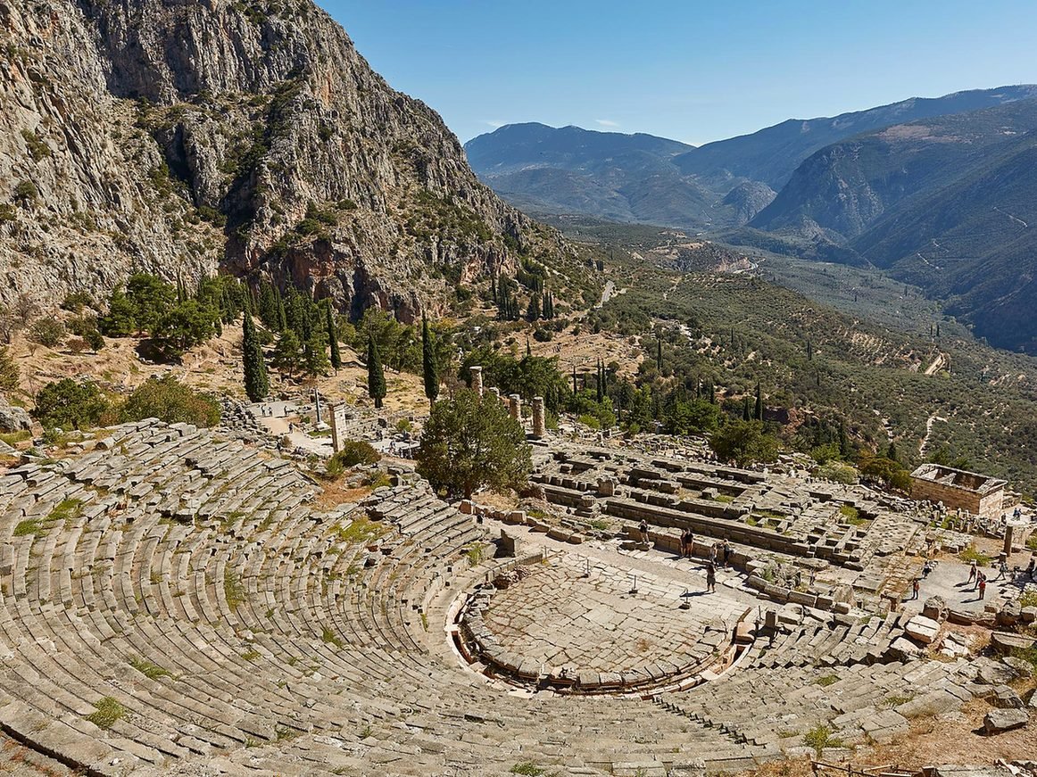 Arquibancadas de pedra do Teatro Antigo de Delfos vistas do alto, com o Templo de Apolo, o Tesouro dos Atenienses e o vale abaixo