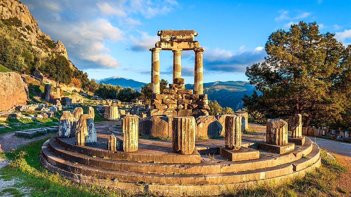 Tholos de Atena Pronaia em Delfos ao entardecer, com tr&ecirc;s colunas d&oacute;ricas reerguidas e montanhas ao fundo