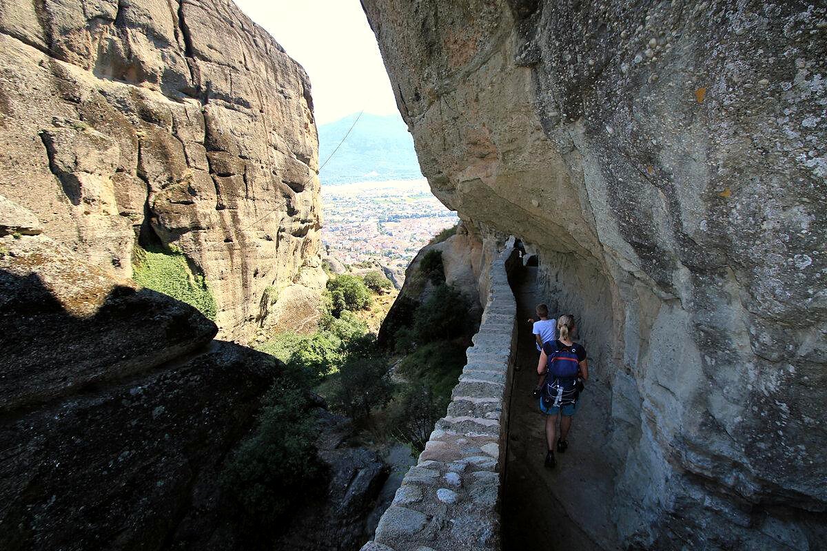 Turistas caminhando por trilha estreita entre os paredões de rocha de Meteora, com Kalambaka ao fundo